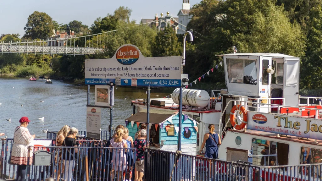 River cruise boat at a dock on the River Dee with passengers and a sign promoting Chester Boat tours.