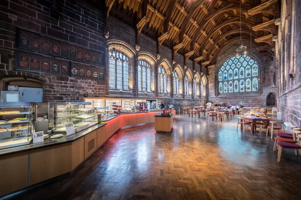 A large, open hall with high wooden arches, stained glass windows, and rows of tables and chairs. A cafeteria-style serving area is on the left side.