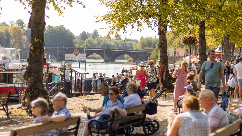 Crowded River Dee promenade with people relaxing on benches, walking, and enjoying the view of the river and a distant bridge.