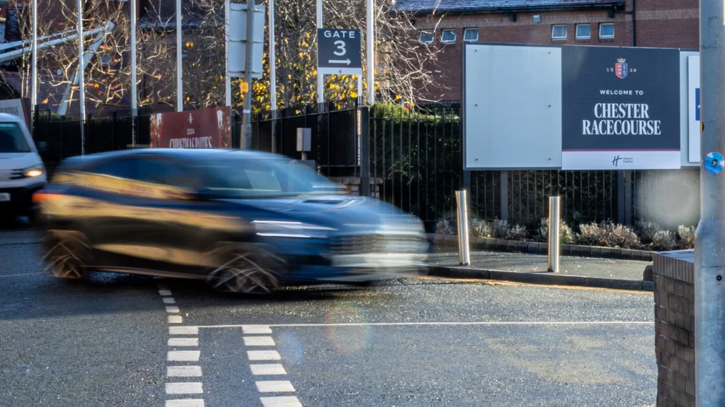 A blurred car drives past Chester Racecourse entrance, sign visible on the right.