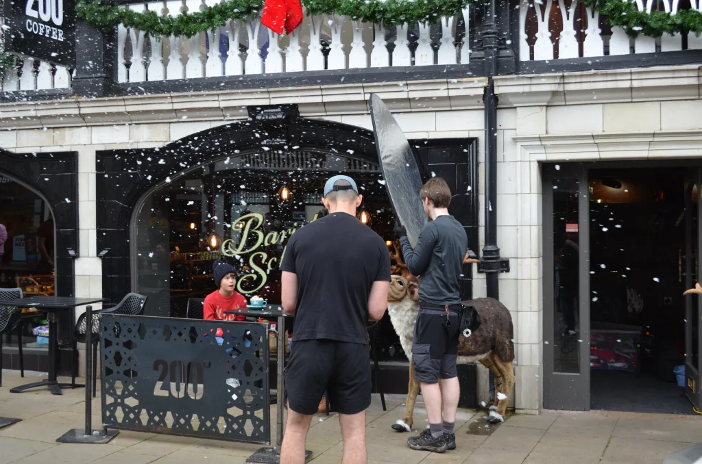 Two photographers outside a restaurant taking photographs of a boy with hot chocolate and a reindeer.