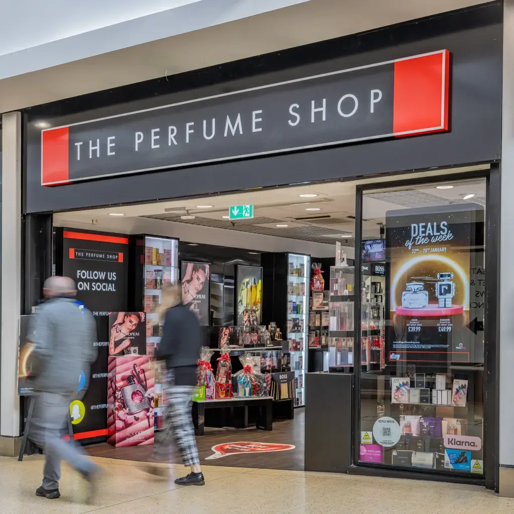 Entrance of "The Perfume Shop," featuring a variety of perfumes and promotional displays, with shoppers walking by.