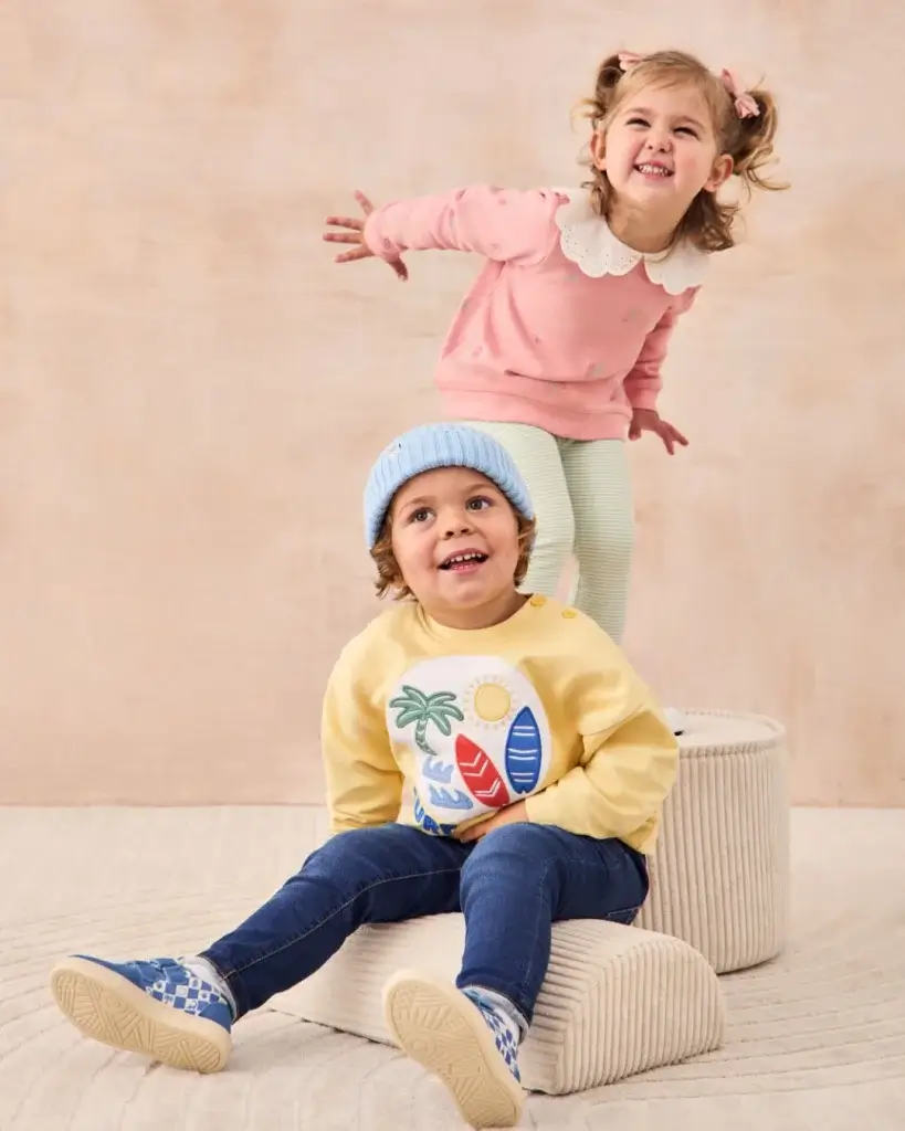 Young Girl smiling in pink and white trainers, white trousers and pink top with white colour and young boy in blue trainers, blue jeans, yellow jumper and light blue hat