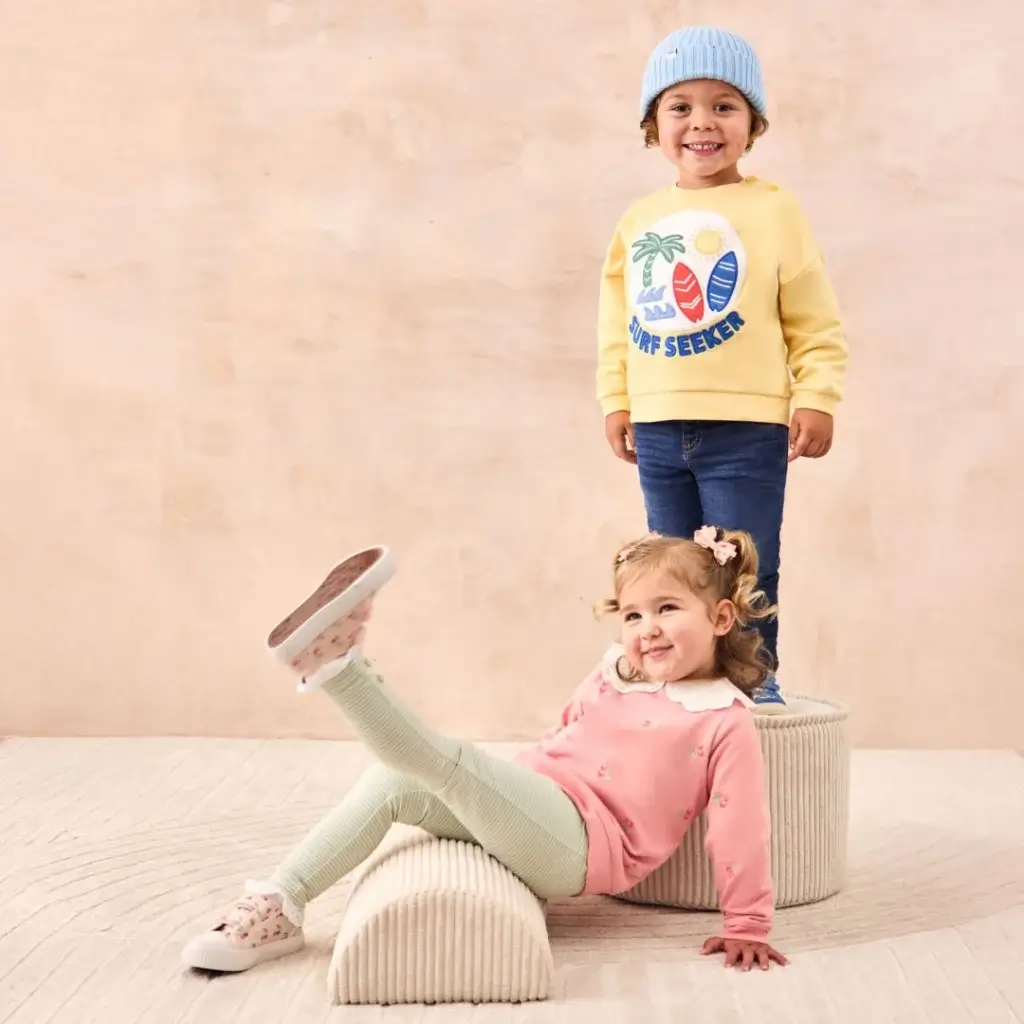 Young Girl smiling in pink and white trainers, white trousers and pink top with white colour and young boy in blue trainers, blue jeans, yellow jumper and light blue hat