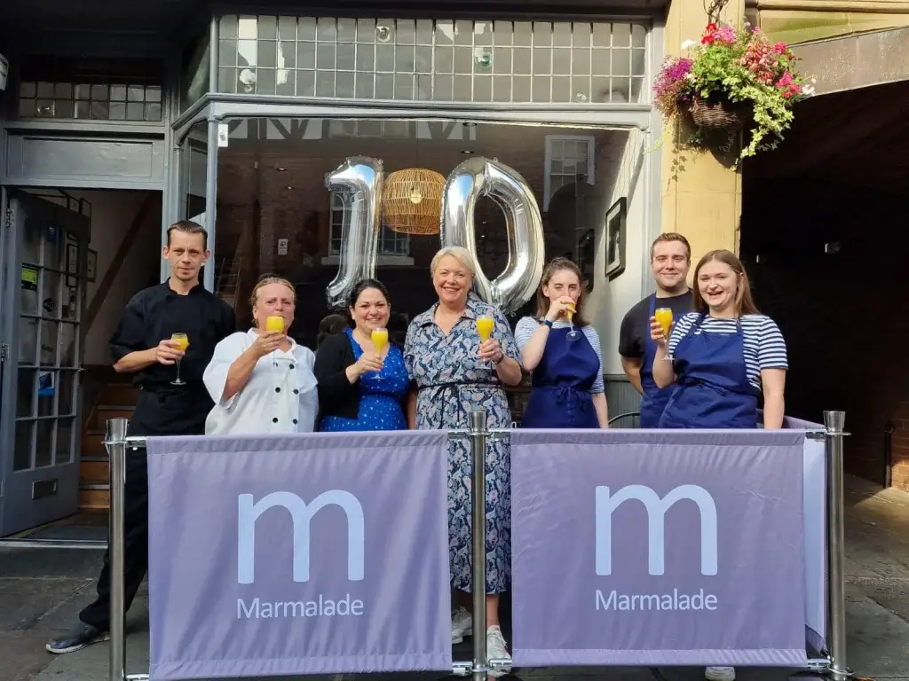 A group of people standing outside of a cafe with balloons shaped in the number 10 holding glasses of Prosecco and orange juice