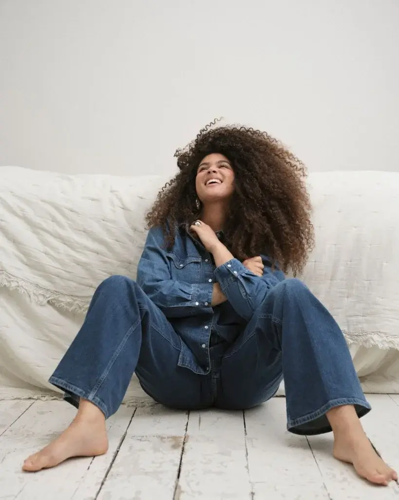 a woman with lots of hair sitting on the ground bare foot in a blue denim onesie