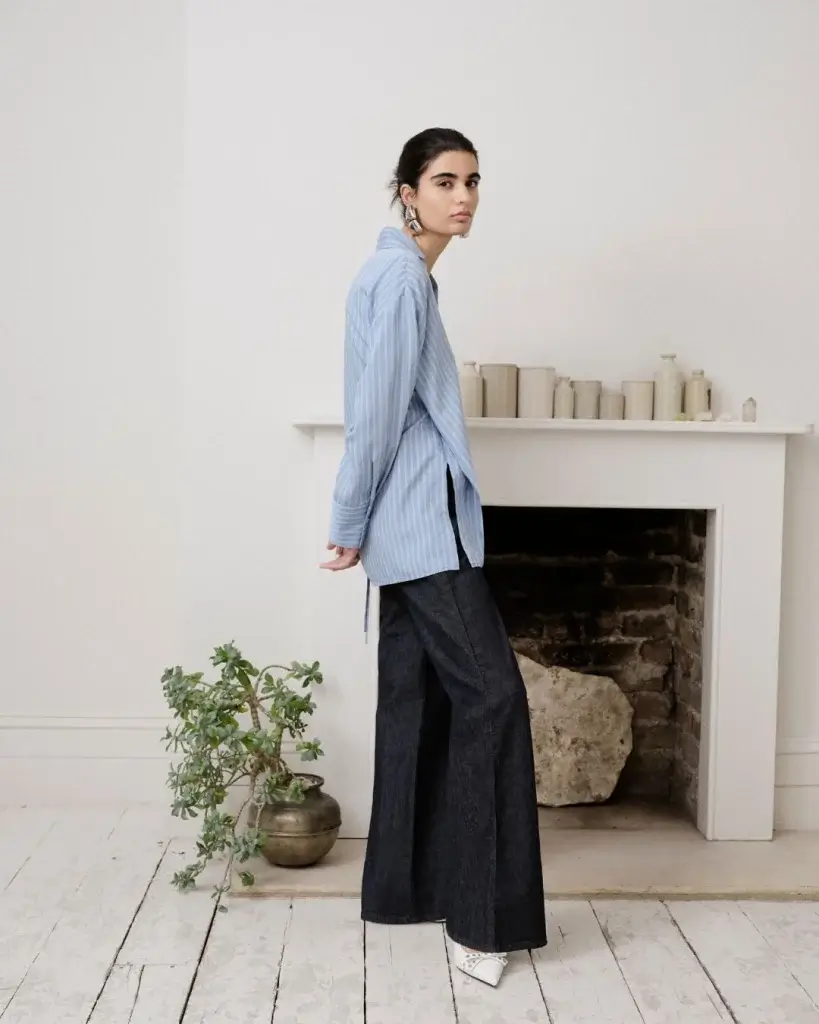 a woman standing up in a blue shit and black trousers standing infant of a white marble fireplace and plant