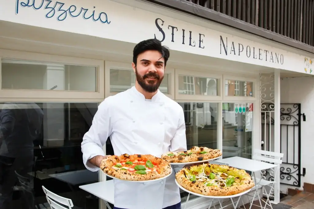 An Italian man holding three pizza outside of a white fronted restaurant