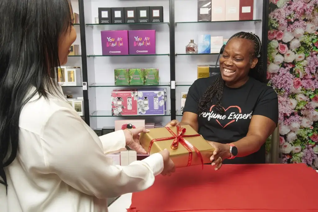 a woman smiling handing of a present to a customer with shelving of perfume in the back