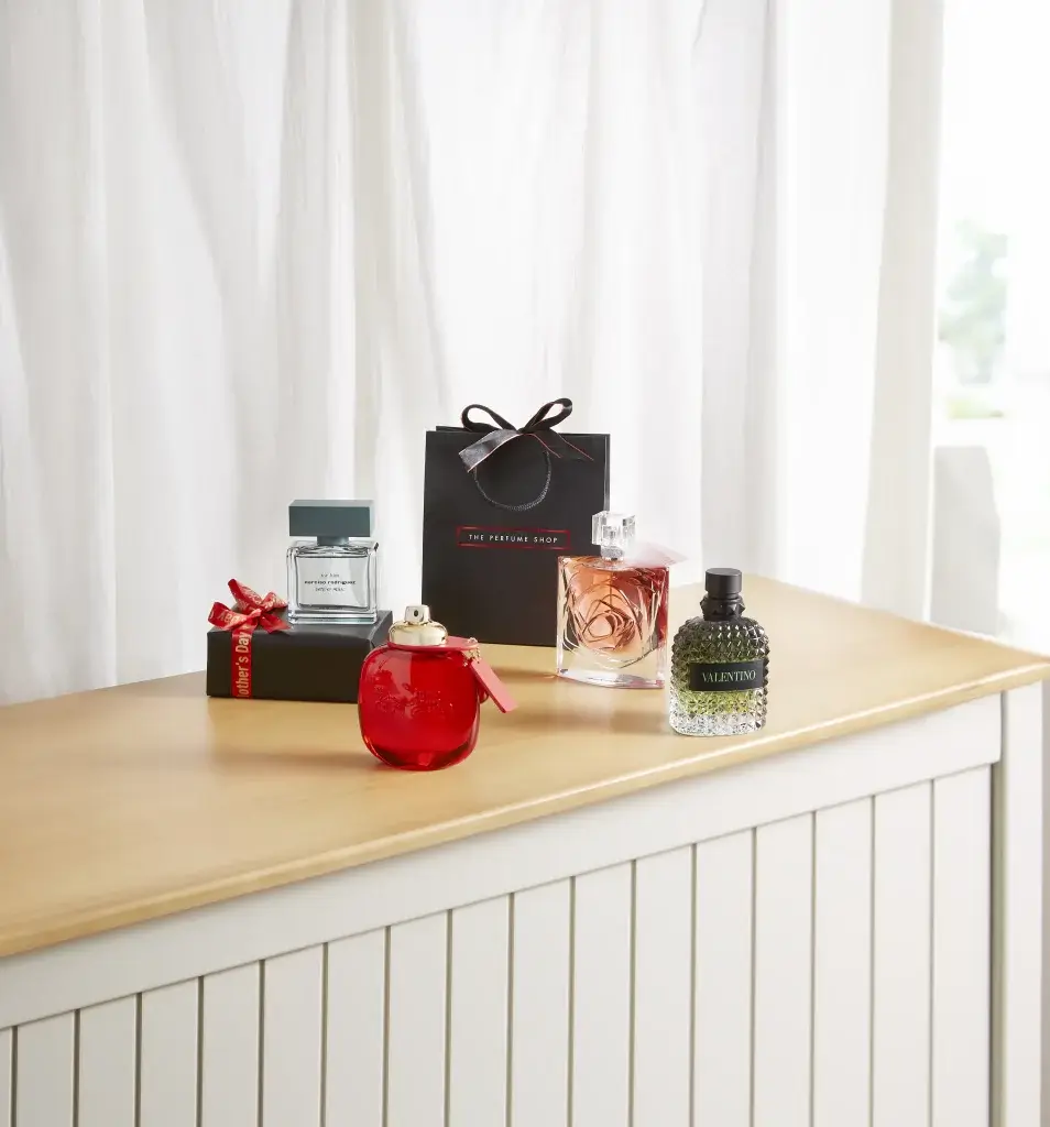 a selection of perfumes on a wooden topped table with a white curtain behind