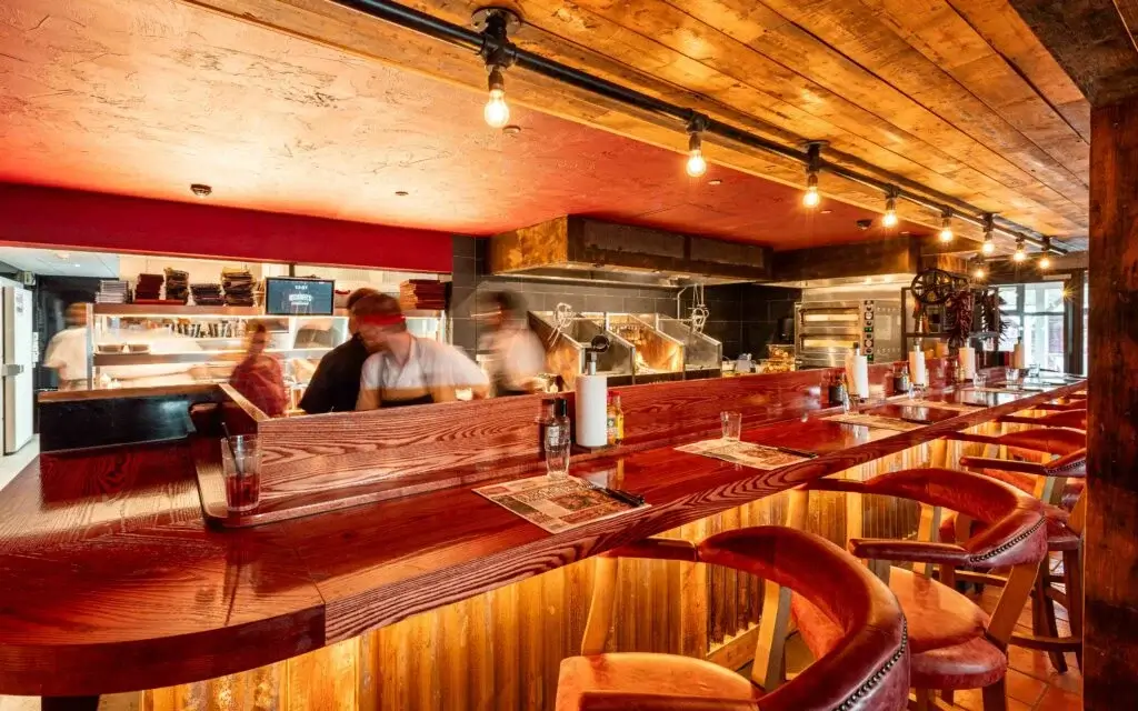 Wooden bar counter with high-backed chairs in a warmly lit restaurant. Staff move in the background near an open kitchen. Glasses and menus are placed on the counter.