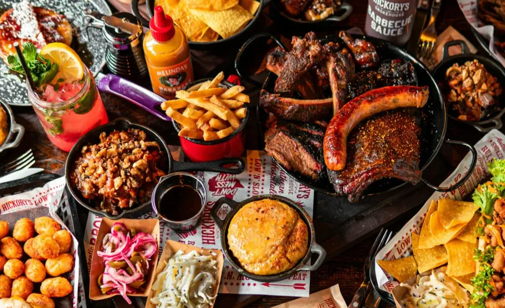 A vibrant spread of barbecue meats, sides, and drinks, featuring ribs, sausage, cornbread, fries, and a colorful cocktail.