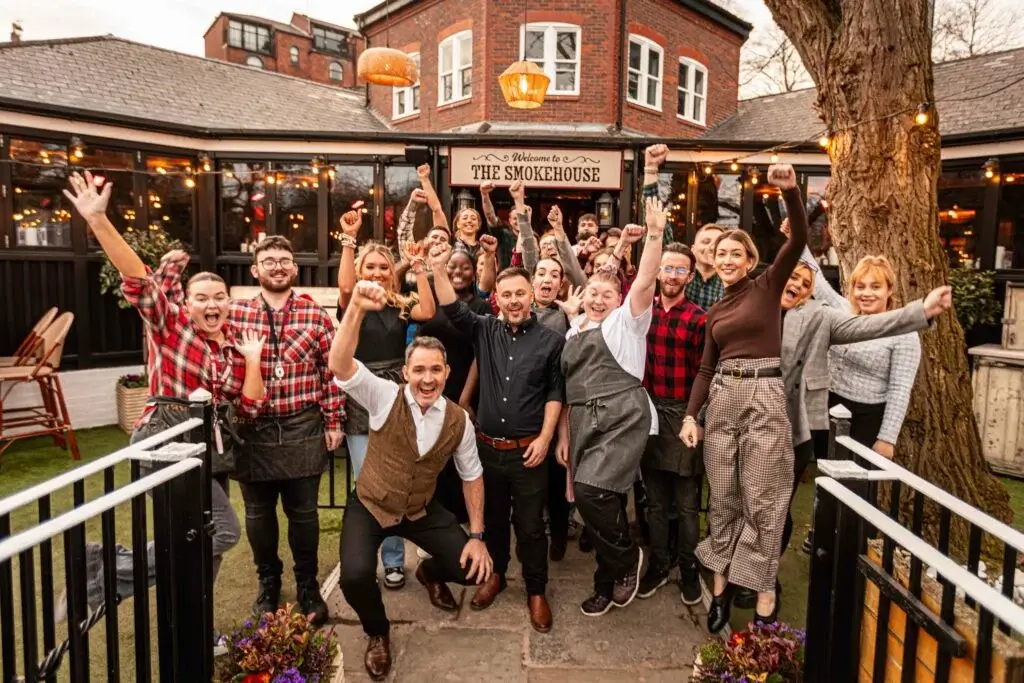 A lively group of staff at "The Smokehouse" celebrates outdoors, posing joyfully with raised hands in a vibrant restaurant setting.