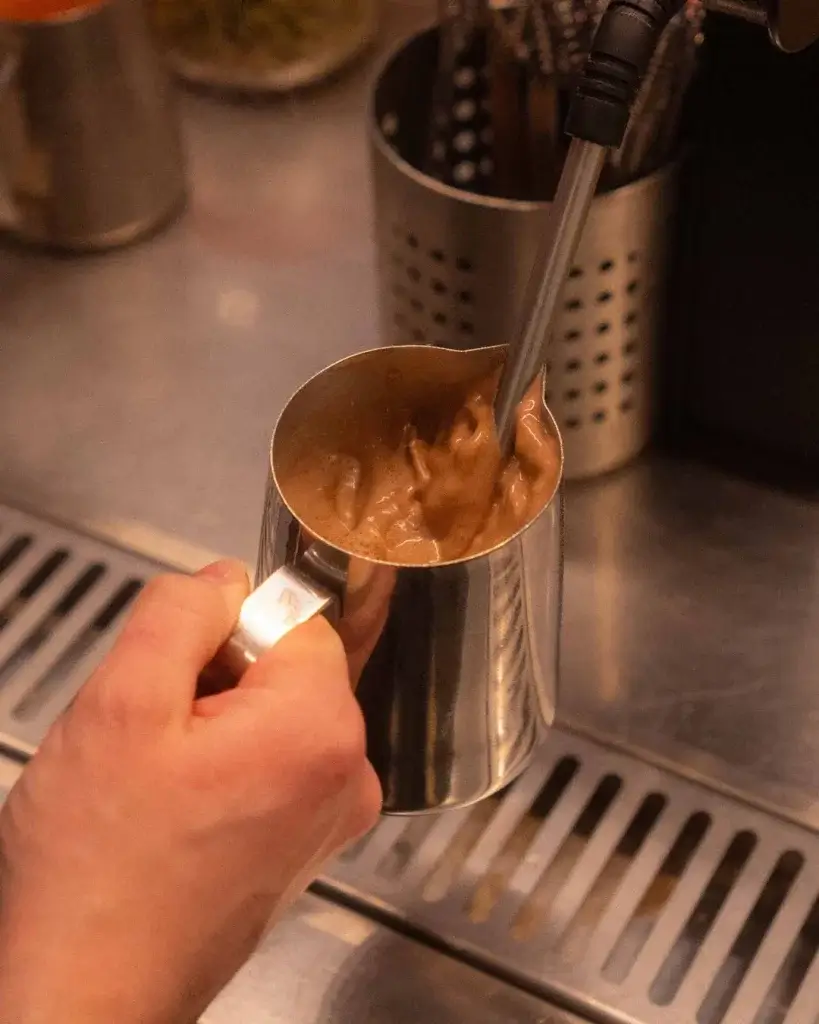 A barista holds a metal pitcher with frothed chocolate, using a steam wand to create creamy foam on a stainless steel counter.