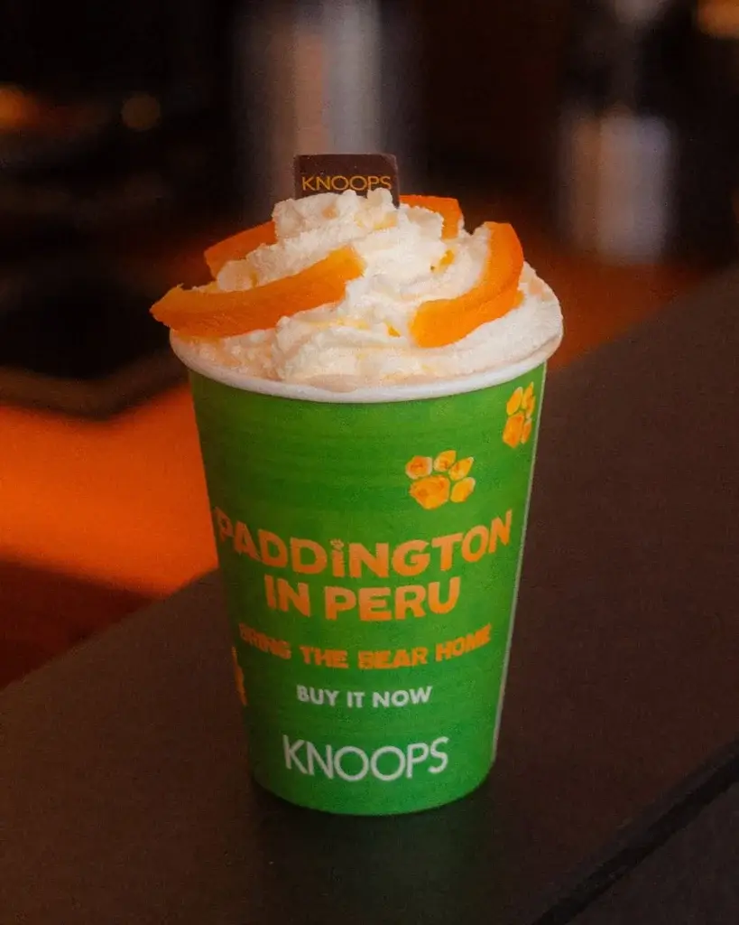 A green cup from KNOOPS featuring "Paddington in Peru" with whipped cream and orange slices on top, promoting a special drink.