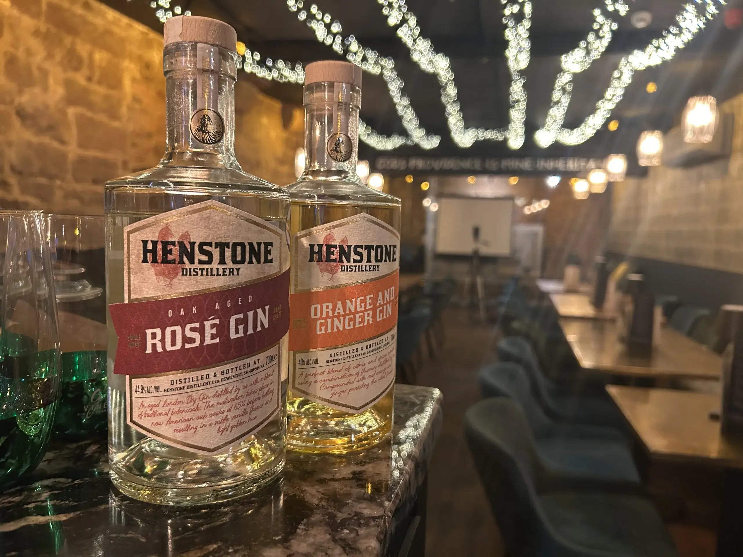 Two bottles of Henstone Distillery gin, one Rose and one Orange & Ginger, displayed in a cozy, twinkling light-filled venue.