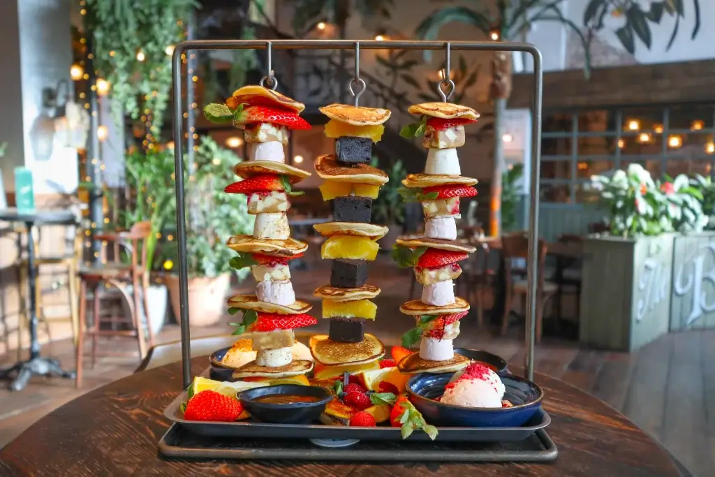 A colorful stack of mini pancakes, fruits, and desserts displayed on a metal stand, served with dipping sauces in bowls.