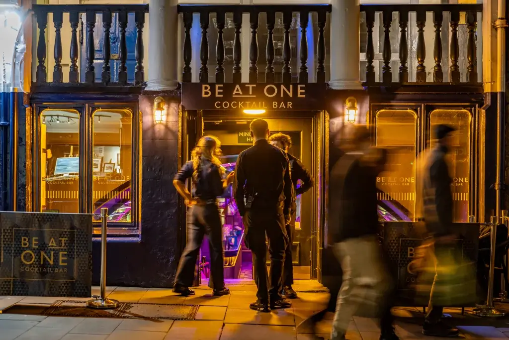 people huddled outside of a bar at night with yellow lighting and purple lighting coming from inside, words be at one can be seen above