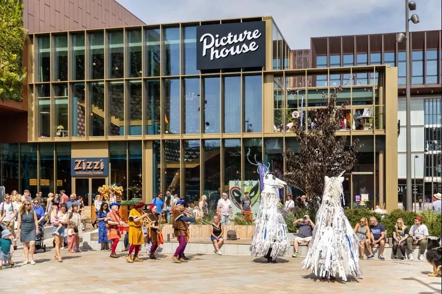 A lively street scene in front of the Picture House, featuring performers in costumes and a crowd enjoying the festivities.