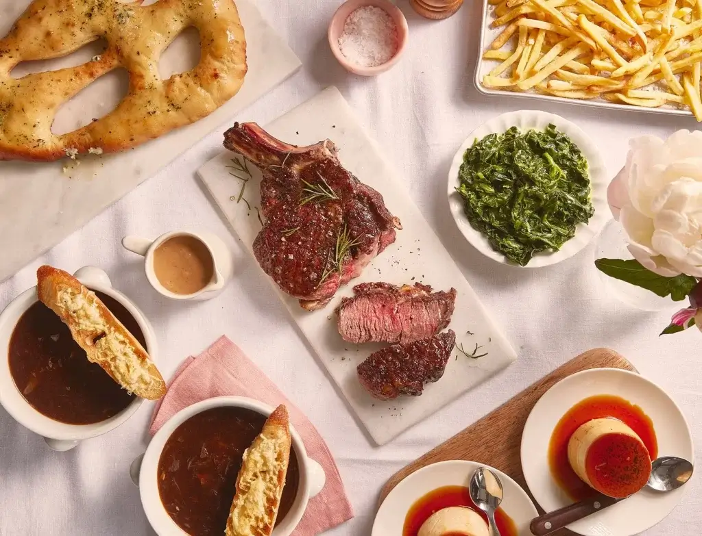 A feast featuring a grilled ribeye steak, sautéed greens, French fries, two bowls of soup with bread, and panna cotta desserts.