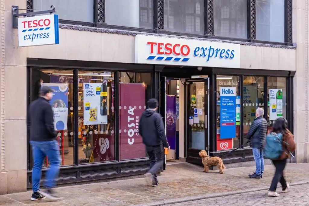 People walk past a Tesco Express store with displays for Costa Coffee and promotional signs, while a dog waits by the entrance.