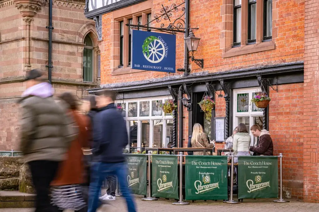 A bustling exterior of a brick bar and restaurant, with patrons seated outside and a hanging sign that reads "The Coach House."